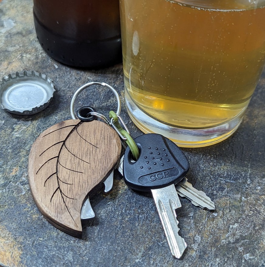 Image of keys on a wooden leaf bottle opener keychain next to a top from a bottle the lower section of a bottle and the lower section of a cup filled with yellow liquid.