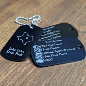 Black military type dog tags with the outline of a state and state park name on one side and a list of it's top trails on the other. The list has boxes to the right of the trail name that you can check off as your complete them.