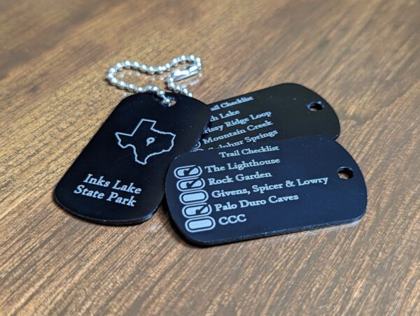 Black military type dog tags with the outline of a state and state park name on one side and a list of it's top trails on the other. The list has boxes to the right of the trail name that you can check off as your complete them.